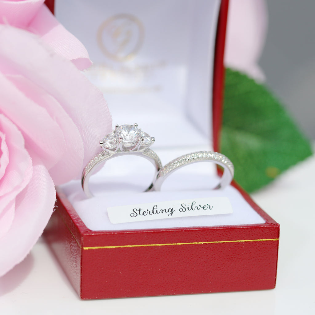 Sterling silver three-stone engagement ring with matching pavé wedding band, displayed in a red ring box with a pink rose and green leaf accent.