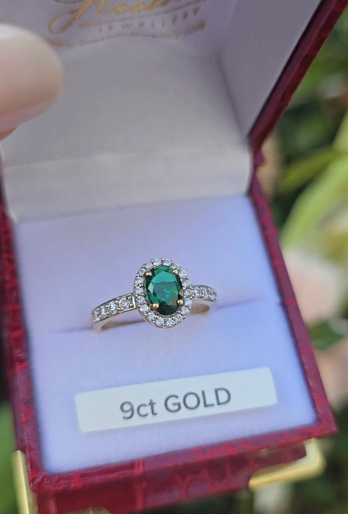 Close-up of a 9ct gold ring with an oval-cut emerald centre stone, surrounded by a halo of cubic zirconia, displayed in a red ring box.