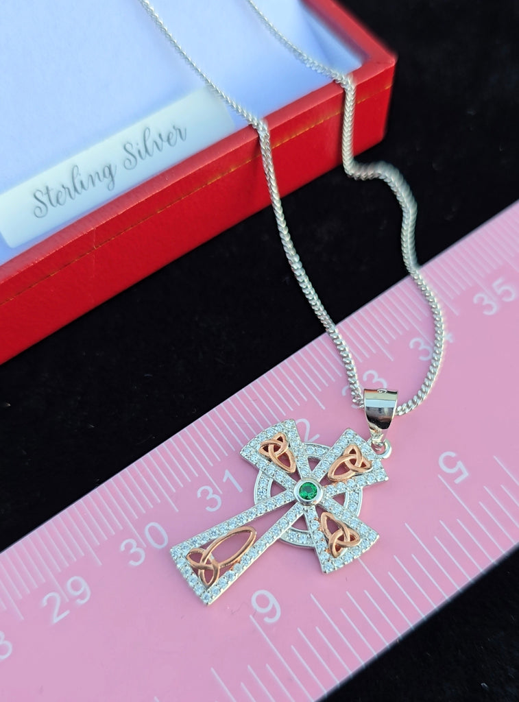 Celtic cross pendant with emerald center placed against a pink ruler for scale, showing dimensions of the piece