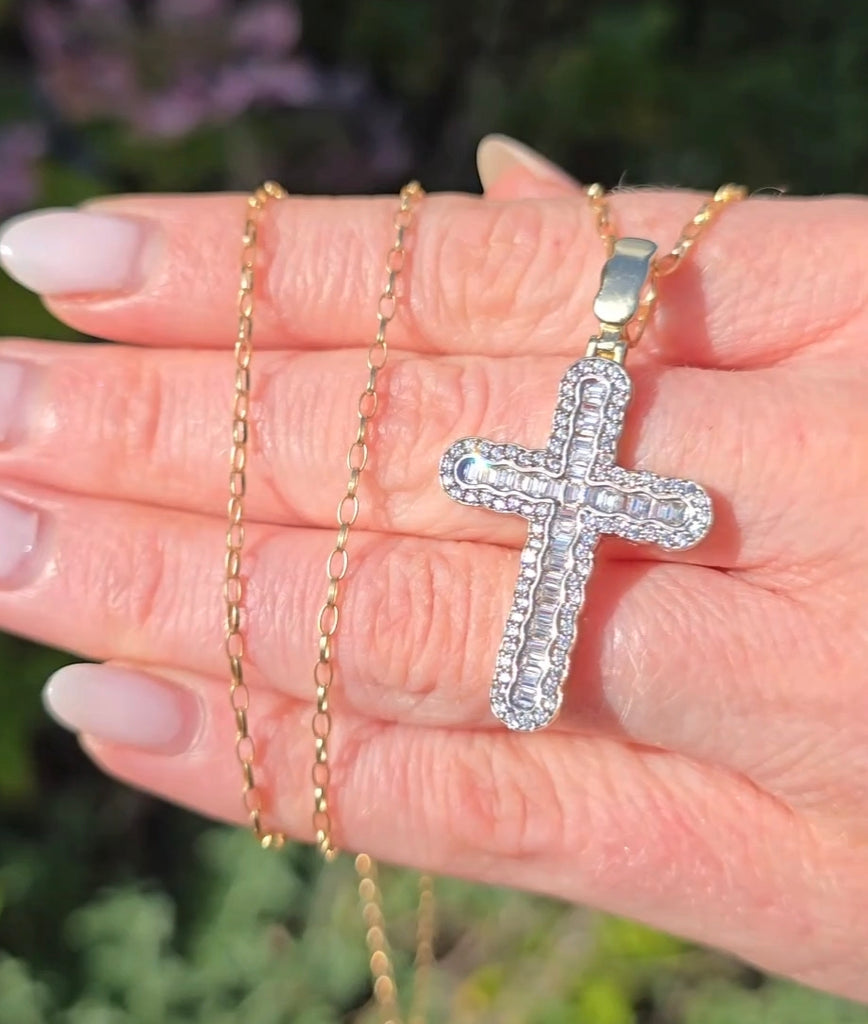 Hand holding 9ct gold cross pendant with baguette and round CZ stones, chain draped over fingers in natural light. Gleeson Jewellers 