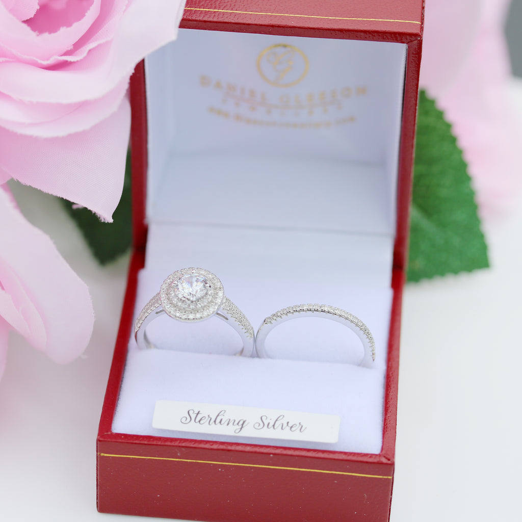 Sterling silver halo engagement and wedding ring set in red presentation box, surrounded by pink flowers and green leaves.