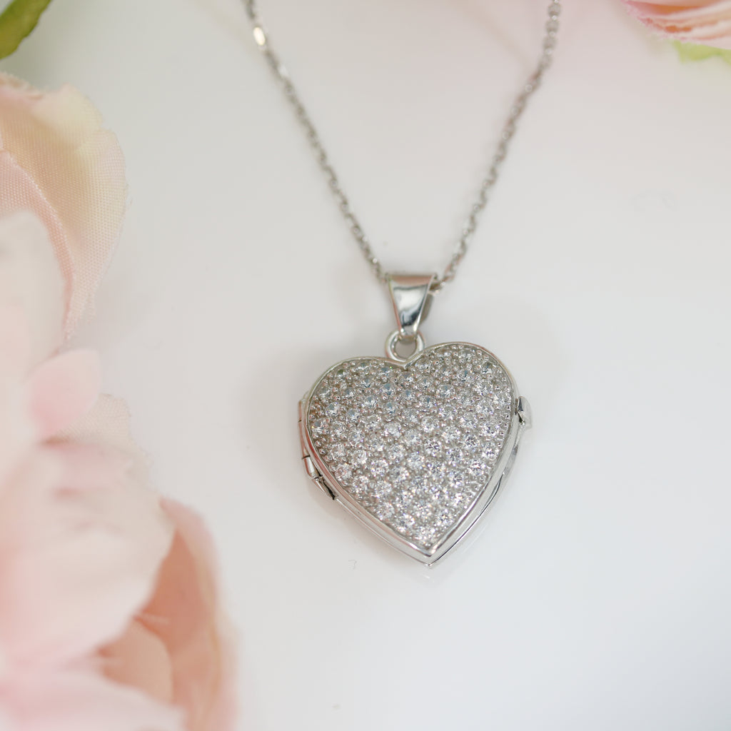 Close-up of a sterling silver heart-shaped locket encrusted with sparkling cubic zirconia stones, hanging from a delicate silver chain against a soft floral background