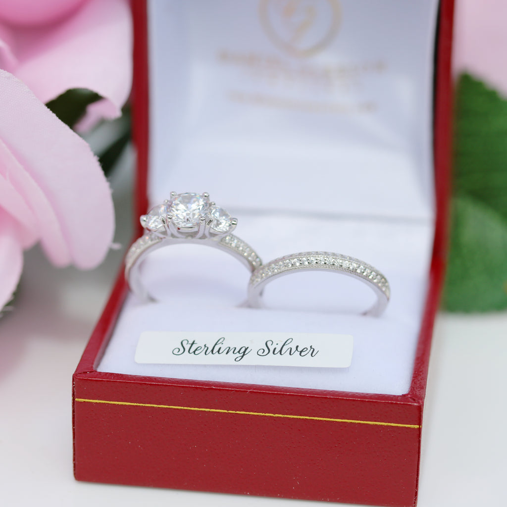 Close-up of sterling silver 3-stone engagement and wedding ring set in red presentation box, white background with pink flower accent.