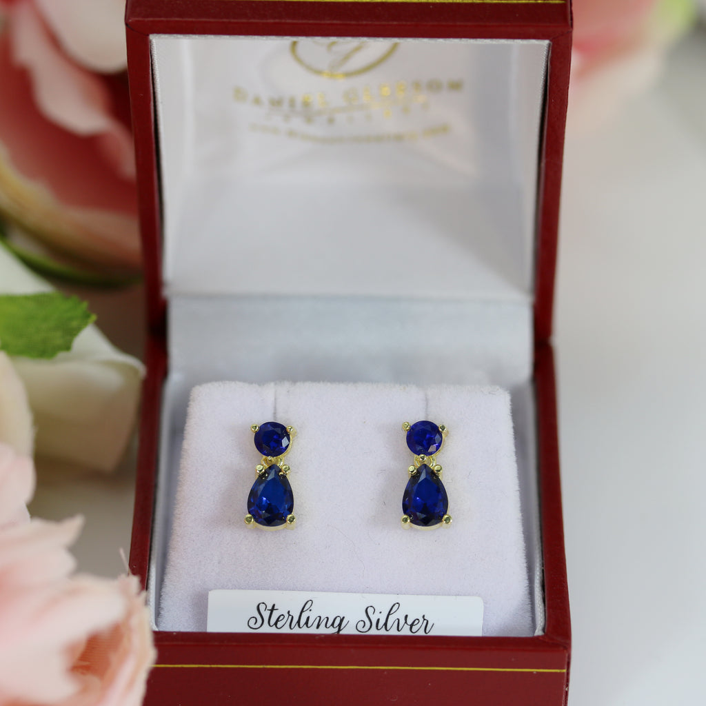 Elegant sapphire CZ petite drop earrings in a classic sterling silver and gold-plated setting, nestled in a red jewellery box with delicate floral accents in the background