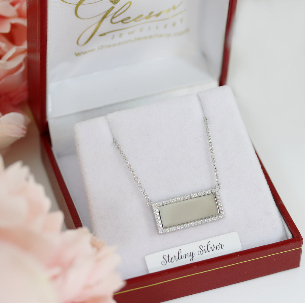 Engravable sterling silver CZ pendant necklace presented in a luxury jewellery box with soft floral accents in the background.