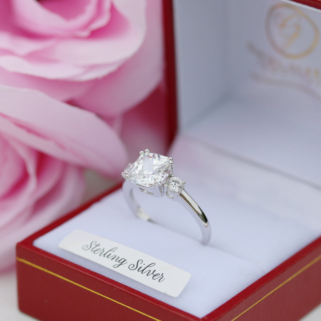  Close-up angled shot of the sterling silver CZ trilogy ring in a red ring box, surrounded by delicate pink rose petals.