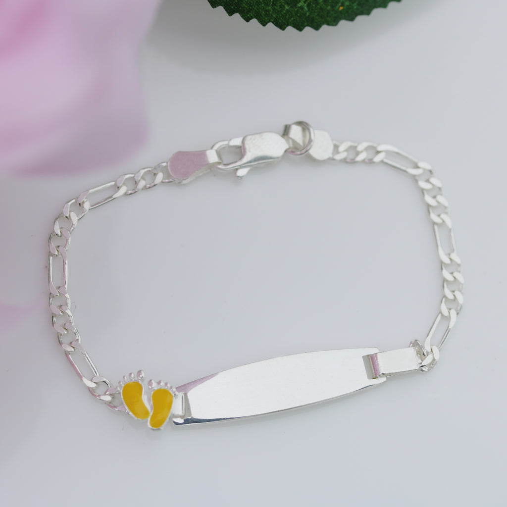 Sterling silver 6.5-inch children’s Figaro ID bracelet with smooth plate and yellow enamel baby feet, displayed on a white background with pink flower detail from Gleeson Jewellers 
