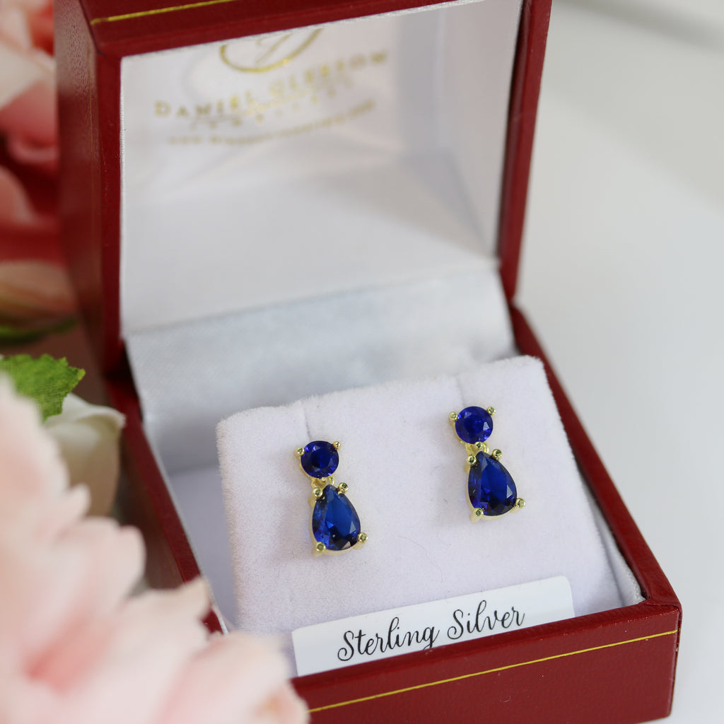 A pair of sterling silver drop earrings featuring deep blue sapphire cubic zirconia stones, elegantly displayed in a red and white jewelry box with soft velvet cushioning